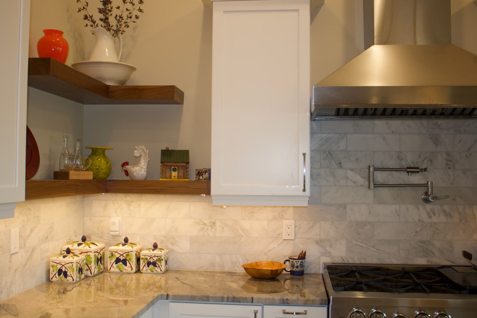 White kitchen corner with marble subway backsplash, walnut floating shelves and stainless hood